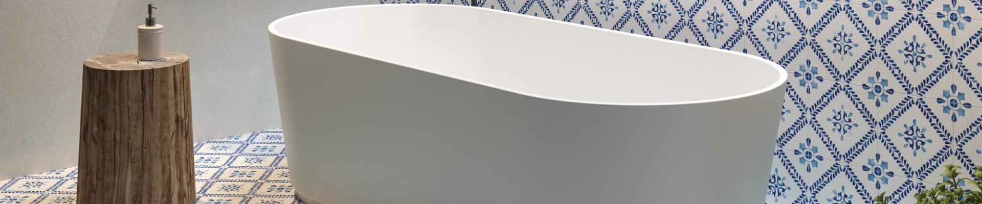 The image shows a modern bathroom setting with a focal point on a standalone bathtub. The bathtub is white with a smooth, curved design, likely made of acrylic or porcelain, and it's positioned in the centre of the room. Behind the tub, there is a section of the wall decorated with blue and white patterned tiles that add a traditional and somewhat Mediterranean touch to the space. The floor is adorned with matching tiles, creating a cohesive and vibrant aesthetic. To the left of the tub, there's a wooden stool that holds a dispenser bottle, possibly soap or lotion. Above the bathtub, a wooden shelf stretches across the wall, holding neatly stacked towels and some bathroom essentials in a wicker basket. On the right side, there is a green plant that introduces a natural element and a pop of colour to the scene. The overall look of the bathroom is clean, serene, and stylish, with a blend of modern fixtures and classic decorative tiles.