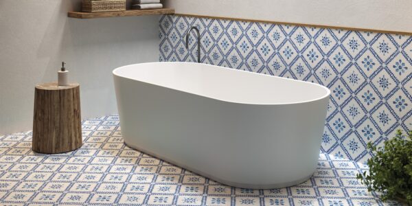 The image shows a modern bathroom setting with a focal point on a standalone bathtub. The bathtub is white with a smooth, curved design, likely made of acrylic or porcelain, and it's positioned in the centre of the room. Behind the tub, there is a section of the wall decorated with blue and white patterned tiles that add a traditional and somewhat Mediterranean touch to the space. The floor is adorned with matching tiles, creating a cohesive and vibrant aesthetic. To the left of the tub, there's a wooden stool that holds a dispenser bottle, possibly soap or lotion. Above the bathtub, a wooden shelf stretches across the wall, holding neatly stacked towels and some bathroom essentials in a wicker basket. On the right side, there is a green plant that introduces a natural element and a pop of colour to the scene. The overall look of the bathroom is clean, serene, and stylish, with a blend of modern fixtures and classic decorative tiles.