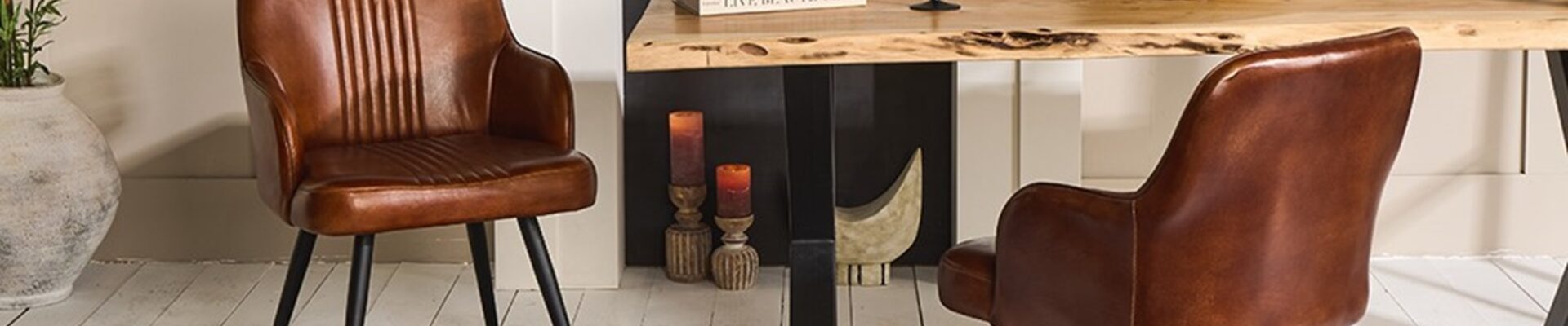 A contemporary dining room featuring a rustic wooden table with live edges, flanked by two brown leather chairs with black metal legs. The table is set against a muted beige wall with a white fireplace mantel. On the table, there's a decorative bowl filled with spheres, a book titled 'Live Beautiful', and two black-stemmed wine glasses. The scene is completed by a textured dark rug underneath and a large potted plant to the left, adding a touch of greenery to the warm-toned space.