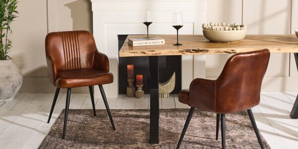 A contemporary dining room featuring a rustic wooden table with live edges, flanked by two brown leather chairs with black metal legs. The table is set against a muted beige wall with a white fireplace mantel. On the table, there's a decorative bowl filled with spheres, a book titled 'Live Beautiful', and two black-stemmed wine glasses. The scene is completed by a textured dark rug underneath and a large potted plant to the left, adding a touch of greenery to the warm-toned space.