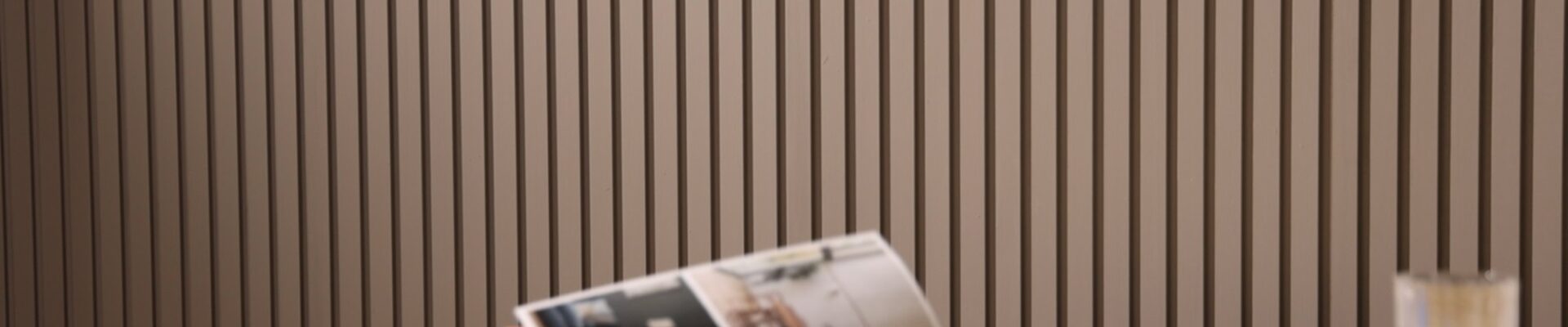 A person reads a magazine at a table. It is an indoor space with a contemporary design, characterized by a slatted wall panelling in the background which adds a textured look to the room. On the table, there's a cup of what might be coffee or tea, a glass of water, and a closed book or journal with a pen on top,