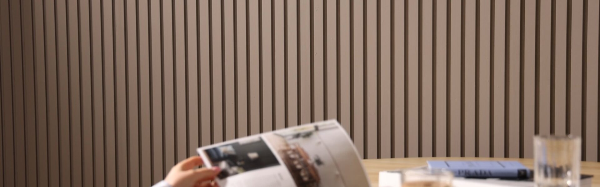 A person reads a magazine at a table. It is an indoor space with a contemporary design, characterized by a slatted wall panelling in the background which adds a textured look to the room. On the table, there's a cup of what might be coffee or tea, a glass of water, and a closed book or journal with a pen on top,