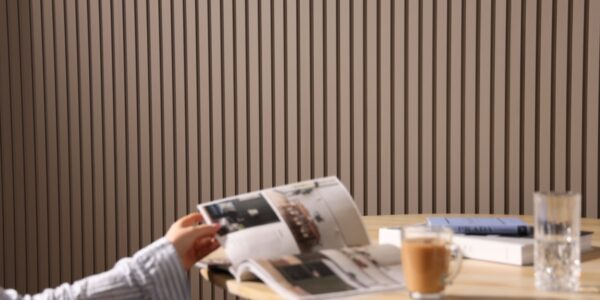 A person reads a magazine at a table. It is an indoor space with a contemporary design, characterized by a slatted wall panelling in the background which adds a textured look to the room. On the table, there's a cup of what might be coffee or tea, a glass of water, and a closed book or journal with a pen on top,