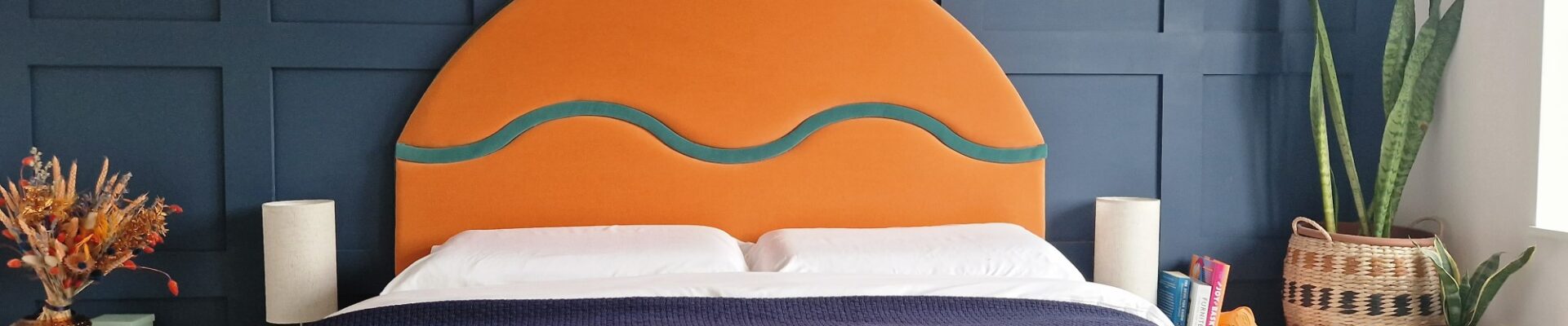 The image shows a bedroom with a contemporary design. The focal point is a large, striking headboard upholstered in a bold orange fabric with a curvilinear teal detail, suggesting a modern and playful style. The headboard sits against a dark blue paneled wall, adding a sense of depth and texture to the room. The bed is covered with a textured blanket featuring abstract shapes in various colors, such as blue, yellow, and orange, which complements the headboard. On either side of the bed are simple, modern white bedside tables, each with a beige lamp, providing a neutral balance to the vibrant colors. There are also decorative elements like a dried flower arrangement in a textured vase, books, and a wicker basket containing green potted plants, enhancing the room's cozy and lived-in feel. The overall composition suggests a well-curated space that blends bold colors and geometric shapes with organic textures.