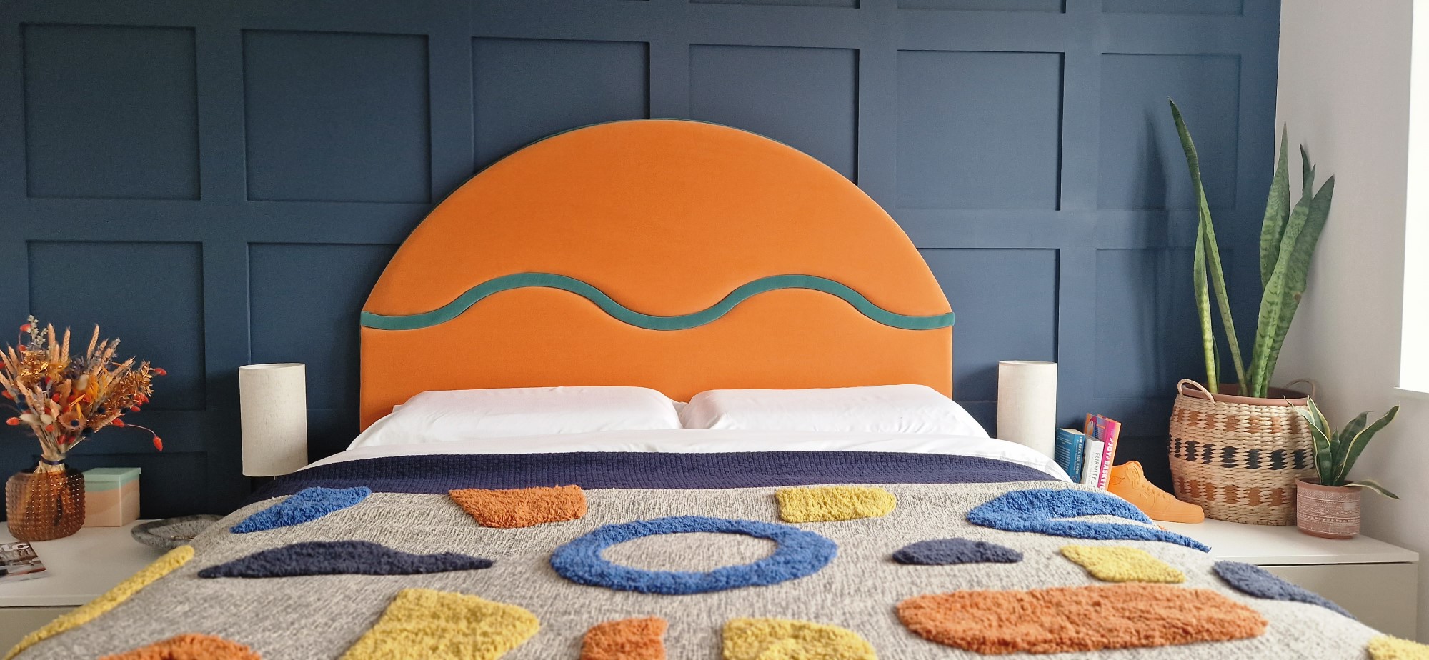 The image shows a bedroom with a contemporary design. The focal point is a large, striking headboard upholstered in a bold orange fabric with a curvilinear teal detail, suggesting a modern and playful style. The headboard sits against a dark blue paneled wall, adding a sense of depth and texture to the room. The bed is covered with a textured blanket featuring abstract shapes in various colors, such as blue, yellow, and orange, which complements the headboard. On either side of the bed are simple, modern white bedside tables, each with a beige lamp, providing a neutral balance to the vibrant colors. There are also decorative elements like a dried flower arrangement in a textured vase, books, and a wicker basket containing green potted plants, enhancing the room's cozy and lived-in feel. The overall composition suggests a well-curated space that blends bold colors and geometric shapes with organic textures.