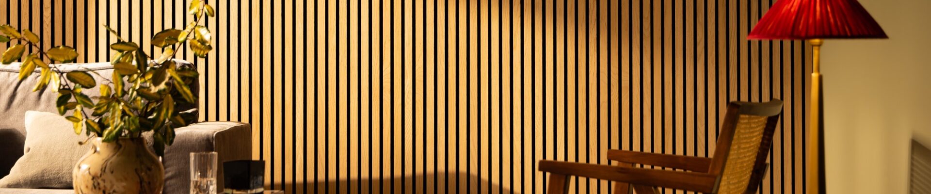 The image shows a living room with a light colored wooden slat wall on the far back wall. There is a gray couch along the left wall, with a light colored wooden end table next to it. A woven basket sits on the floor next to the end table. On the right side of the image is a light colored wooden armchair with a brown leather cushion. A small round coffee table sits in the center of the room, in front of the couch. A black lamp with a white shade sits on the coffee table.
