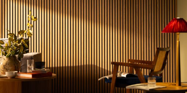 The image shows a living room with a light colored wooden slat wall on the far back wall. There is a gray couch along the left wall, with a light colored wooden end table next to it. A woven basket sits on the floor next to the end table. On the right side of the image is a light colored wooden armchair with a brown leather cushion. A small round coffee table sits in the center of the room, in front of the couch. A black lamp with a white shade sits on the coffee table.
