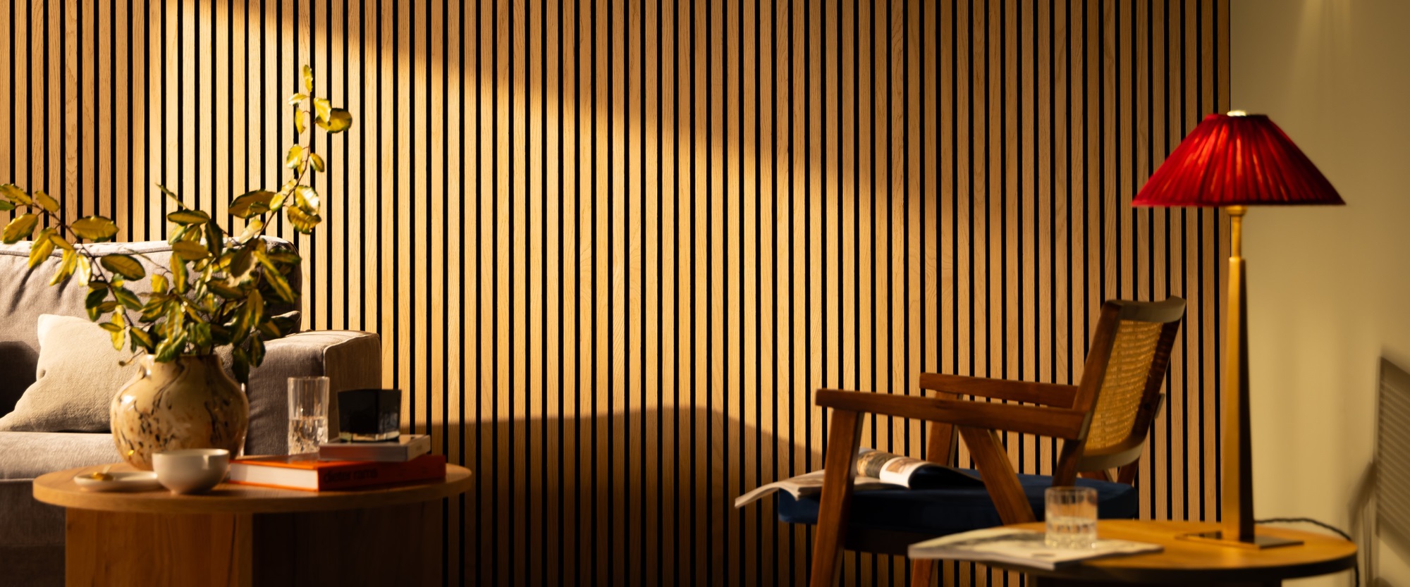 The image shows a living room with a light colored wooden slat wall on the far back wall. There is a gray couch along the left wall, with a light colored wooden end table next to it. A woven basket sits on the floor next to the end table. On the right side of the image is a light colored wooden armchair with a brown leather cushion. A small round coffee table sits in the center of the room, in front of the couch. A black lamp with a white shade sits on the coffee table.
