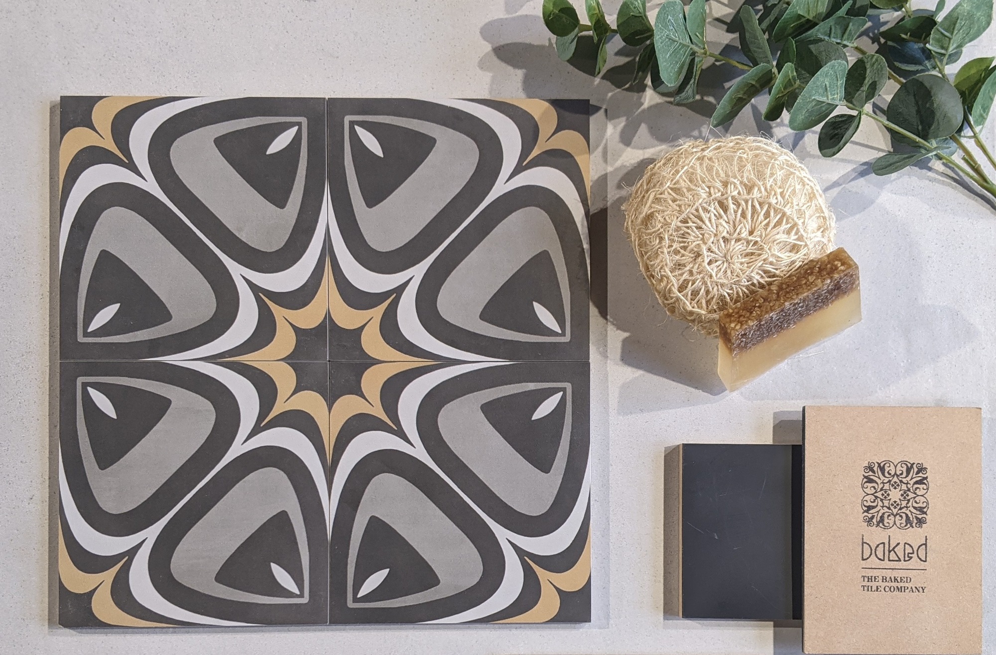 A selection of bathroom tiles in different colours and patterns, with a soap dish and sponge placed on top. The Baked Tile Company logo is visible in the background.