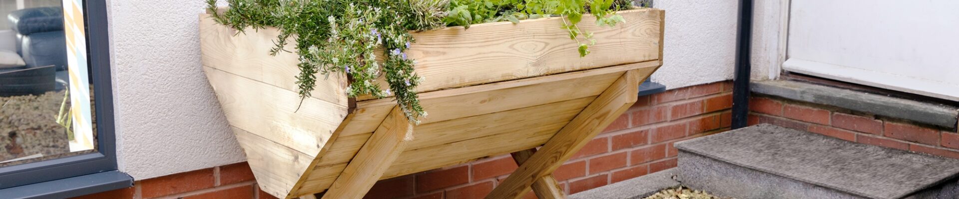 A wooden planter filled with a variety of colourful flowers and plants sits on a gravel surface next to a red brick wall.
