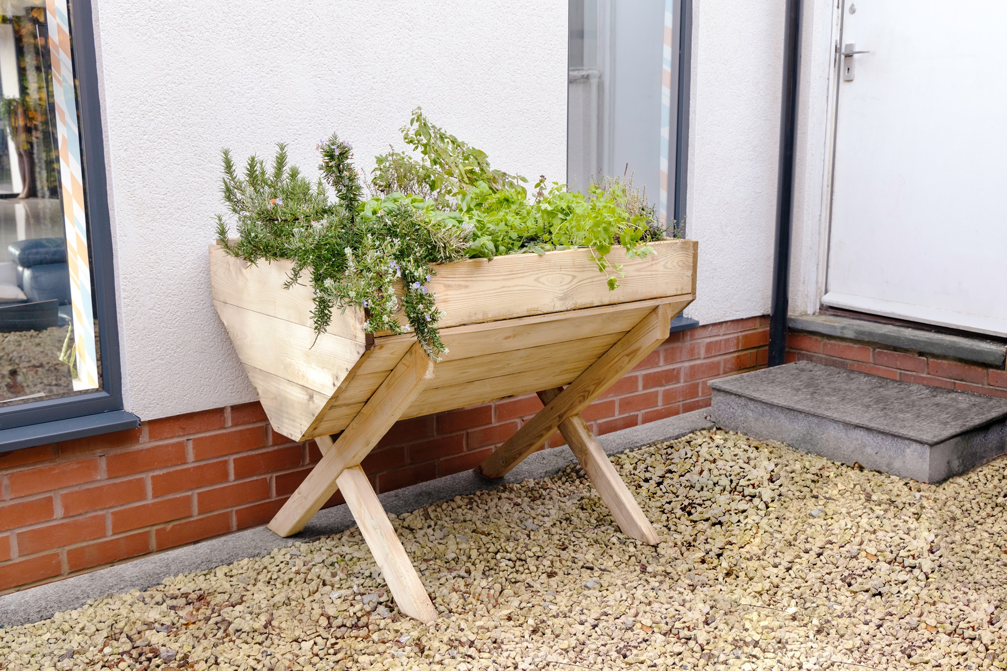 A wooden planter filled with a variety of colourful flowers and plants sits on a gravel surface next to a red brick wall.