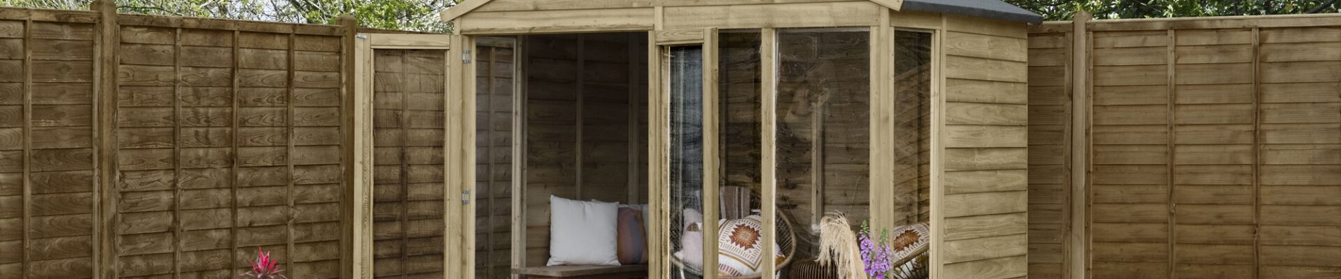 A brown wooden summerhouse is located in a garden. The shed has two doors and four windows. A wooden fence runs along the edge of the garden.