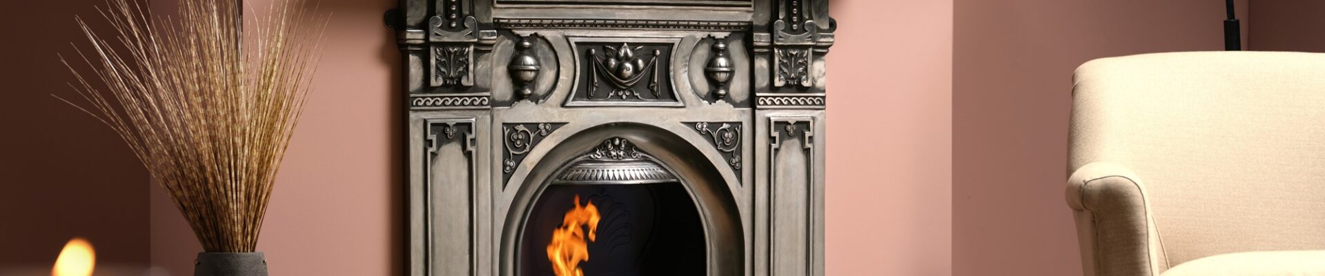 A cast iron fireplace with a fire burning in the grate. The fireplace is in a living room with an armchair next to it.