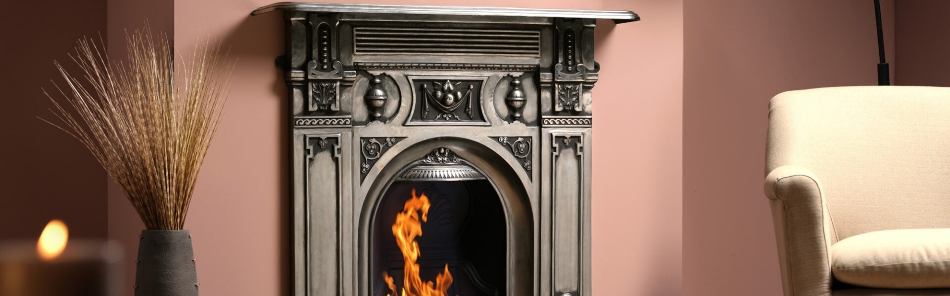 A cast iron fireplace with a fire burning in the grate. The fireplace is in a living room with an armchair next to it.