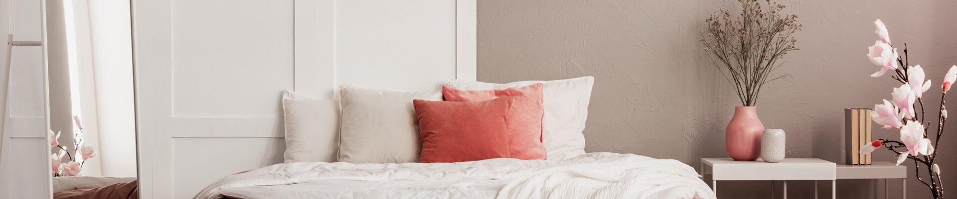 A cosy bedroom with a wooden bed frame, a nightstand with a lamp on top, and a large rectangular mirror hanging on the wall above the nightstand. The bedding is white and the walls are cream coloured.