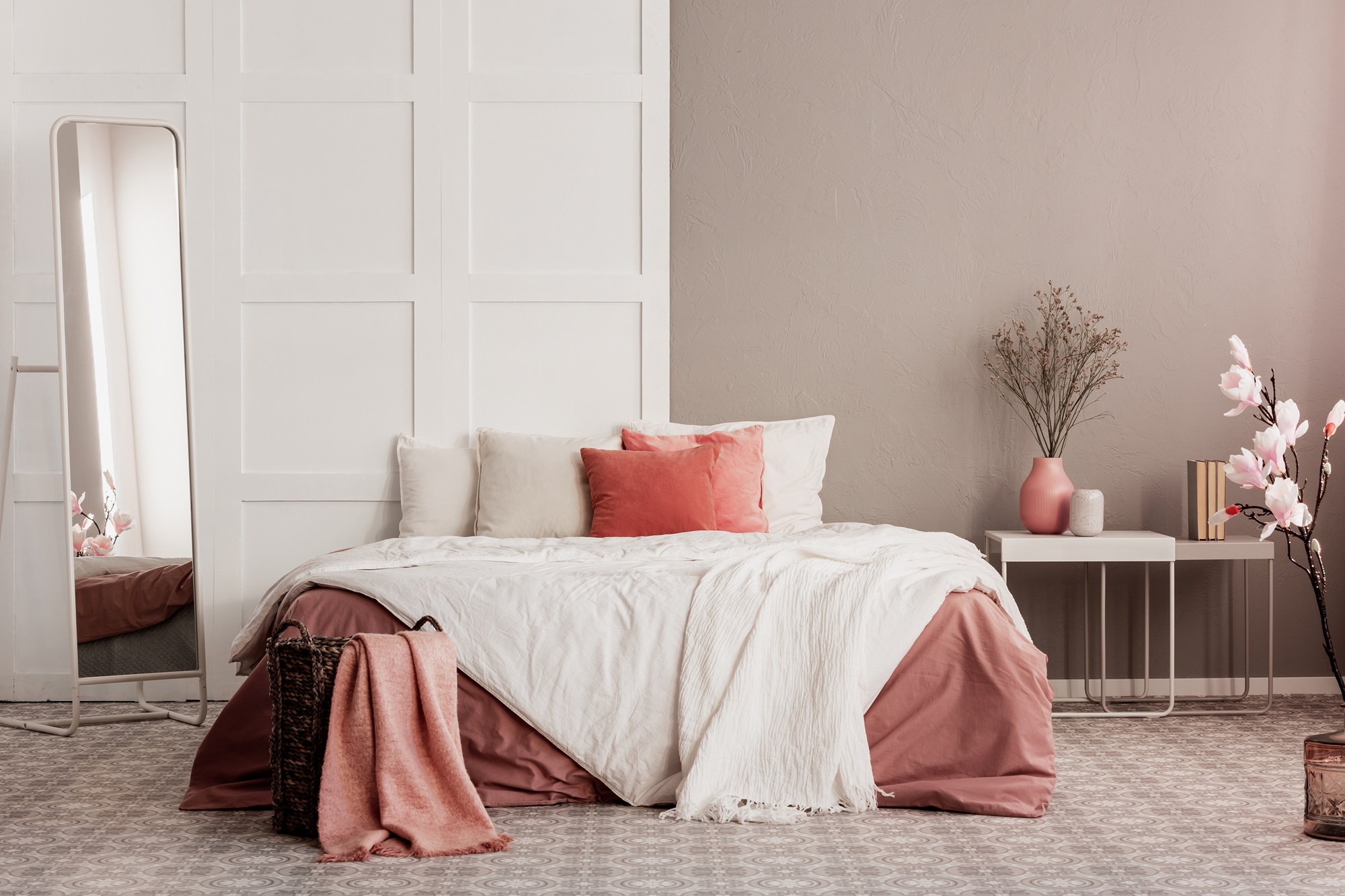 A cosy bedroom with a wooden bed frame, a nightstand with a lamp on top, and a large rectangular mirror hanging on the wall above the nightstand. The bedding is white and the walls are cream coloured.