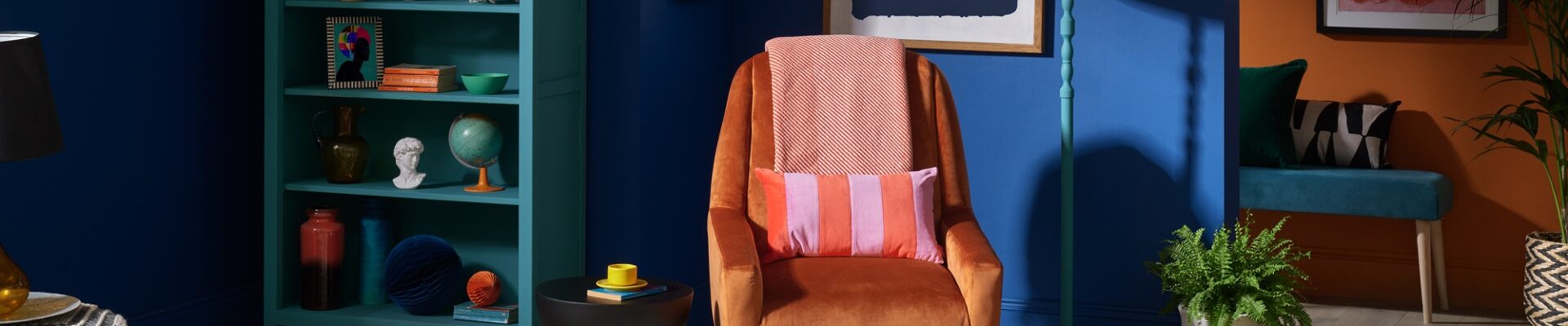 A cosy living room with a wooden armchair upholstered in light fabric sitting on a colourful rug. A wooden bookshelf filled with books is next to the armchair. A table lamp with a white shade sits on a small table beside the armchair.