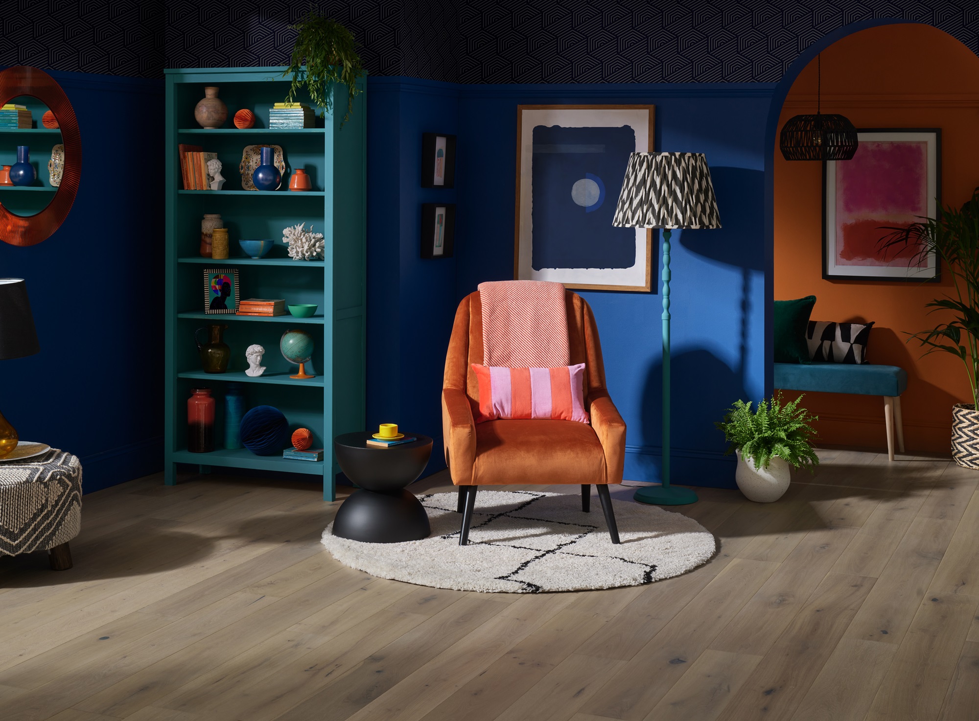 A cosy living room with a wooden armchair upholstered in light fabric sitting on a colourful rug. A wooden bookshelf filled with books is next to the armchair. A table lamp with a white shade sits on a small table beside the armchair.