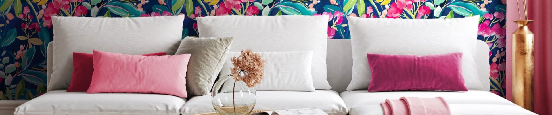 Living room with colourful floral wallpaper on one wall and a pink couch with a round coffee table in front of it. There is a white lamp on the coffee table and a green plant behind the couch.
