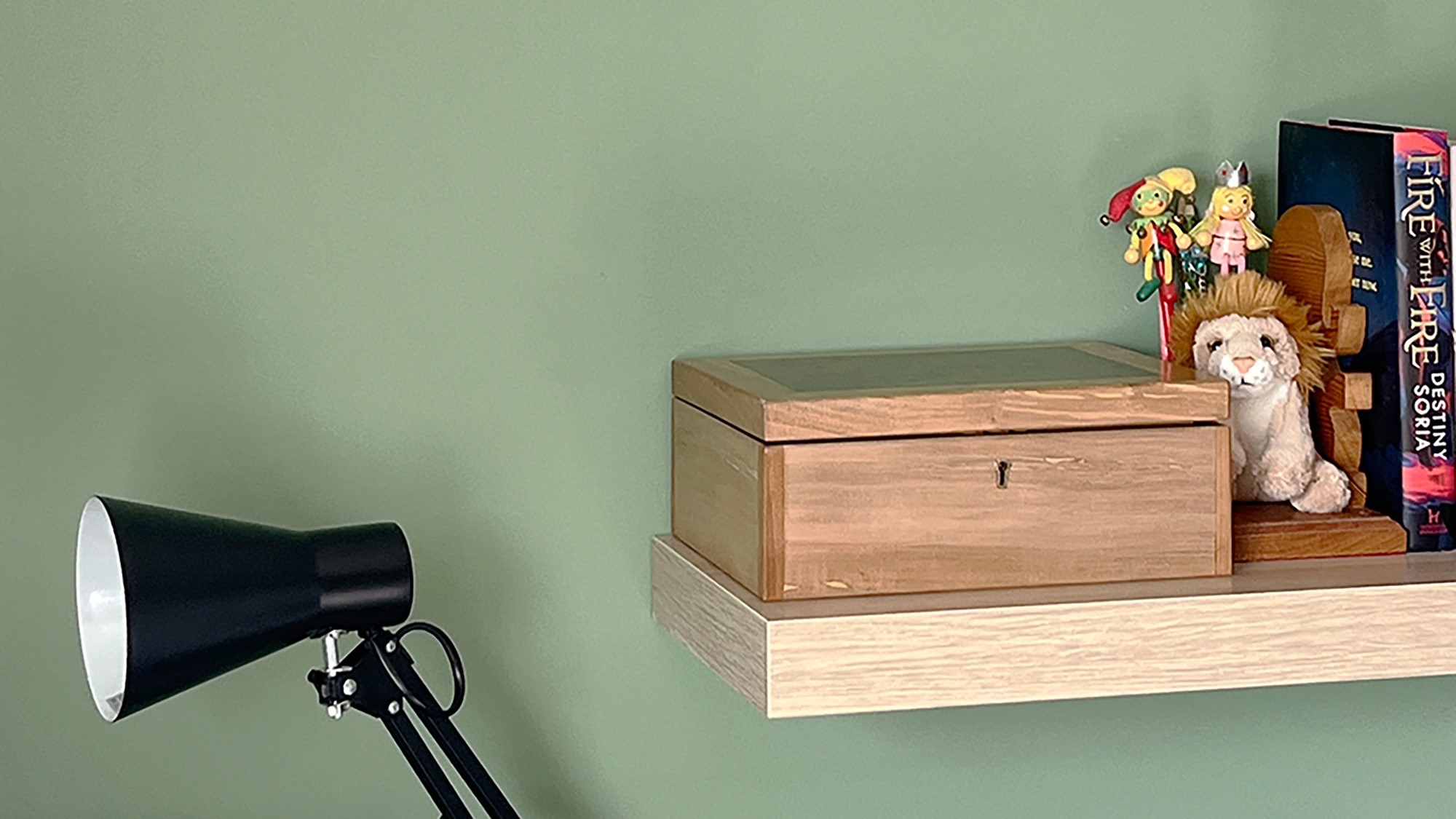 A wooden shelf mounted on a green wall with a decorative box, book and a lamp. The box has the words “Fire With Fire” and “Destiny Soria” written on it. The lamp is a black, metal lamp.