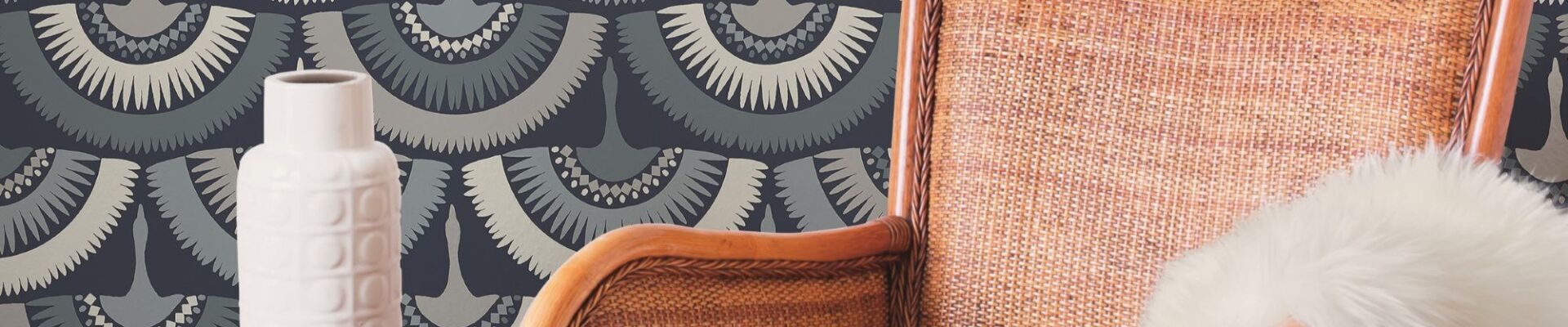 The wallpaper has a blue background with a white and grey feather pattern. The feathers are arranged in a staggered pattern, and they look like they are blowing in the wind. There is a brown armchair in the foreground, and a white vase and book on a side table in the background.