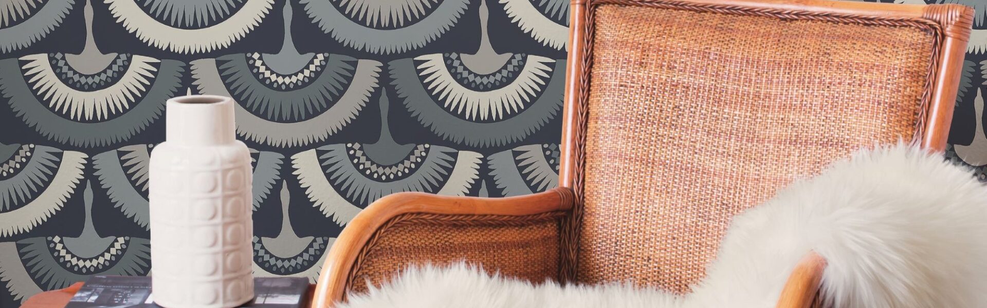 The wallpaper has a blue background with a white and grey feather pattern. The feathers are arranged in a staggered pattern, and they look like they are blowing in the wind. There is a brown armchair in the foreground, and a white vase and book on a side table in the background.