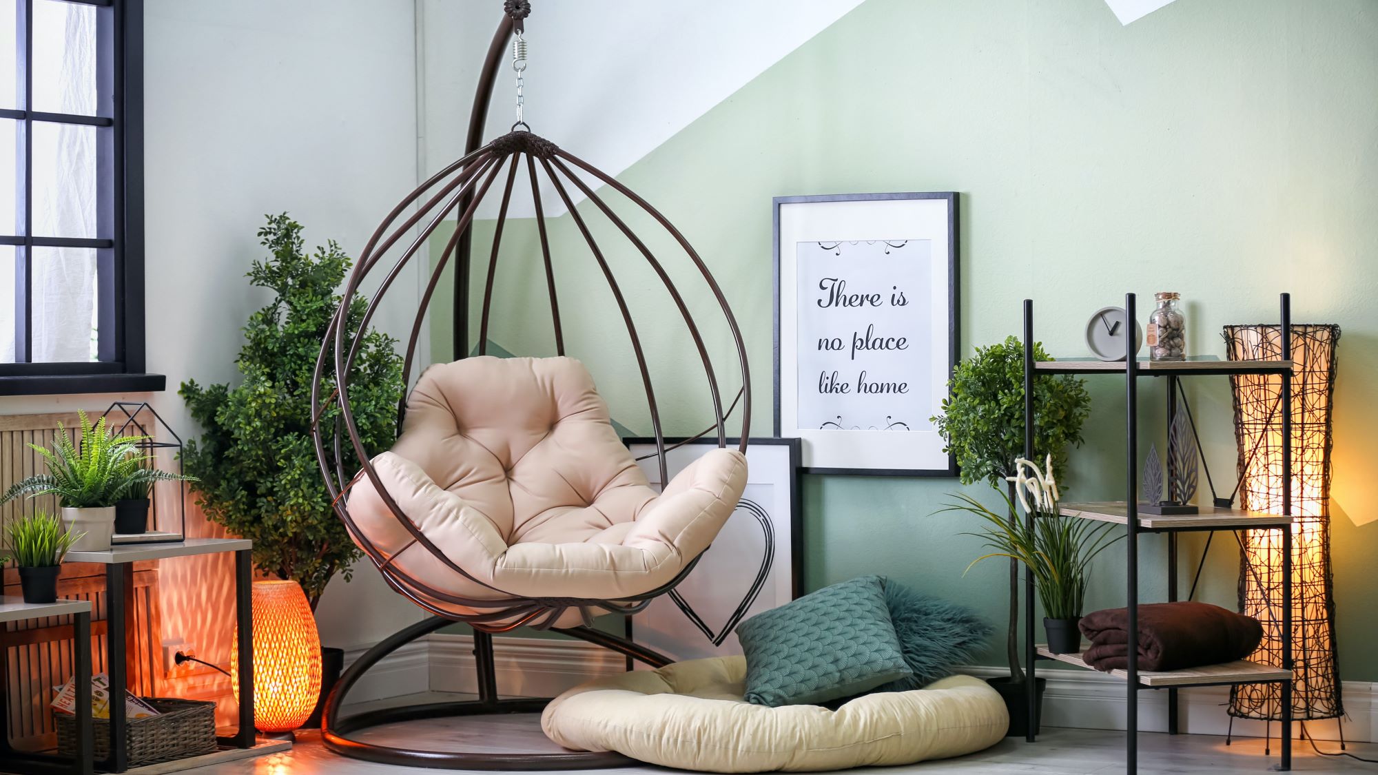 A cosy living room in sage green with a hanging chair in the corner. The chair is made of woven rope and has a cream-coloured cushion. There is a small side table next to the chair with a cup of coffee and a book on it.