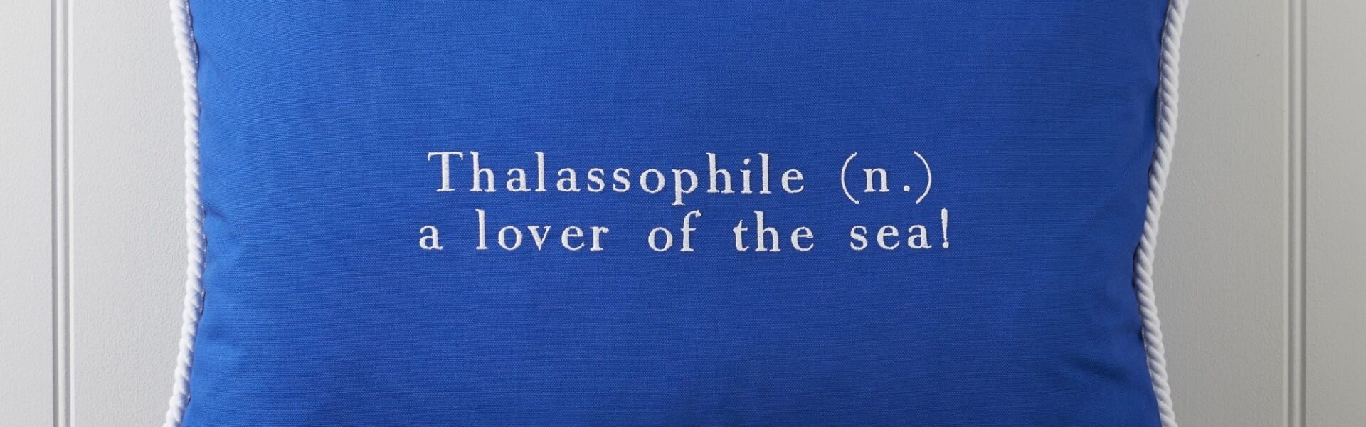 Cushion featuring the word "Thalassophile" embroidered on it in white. The word "Thalassophile" means "a lover of the sea". The cushion also has a white rope trim around the edges.