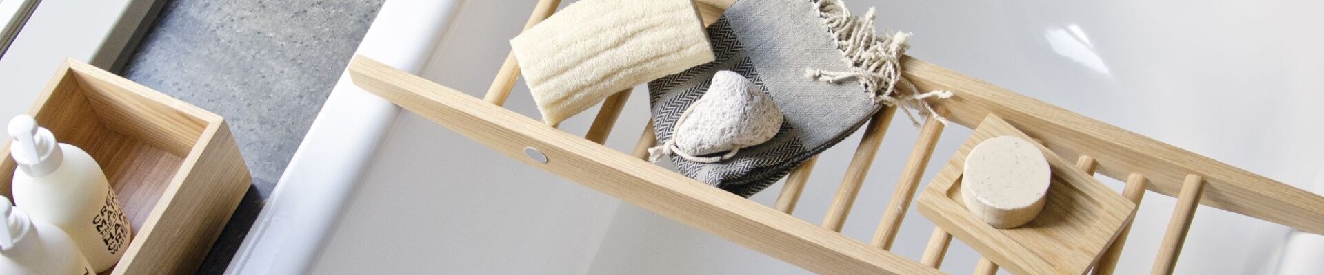 The image shows a Wireworks Bath Bridge in a bathtub. The bath bridge is made of natural oak and has a ladder-like design. It is placed across the bathtub and holds a variety of bath accessories, including a towel, soap, and a loofah. The bathtub is white and has a modern design.