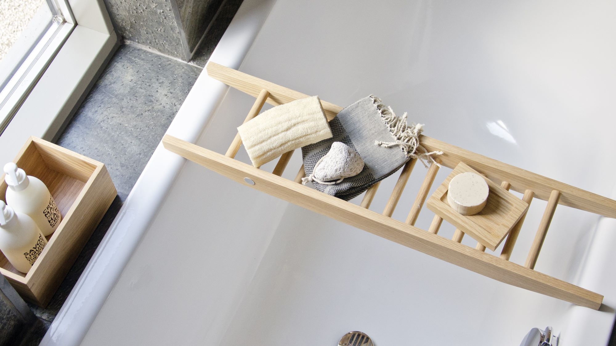 The image shows a Wireworks Bath Bridge in a bathtub. The bath bridge is made of natural oak and has a ladder-like design. It is placed across the bathtub and holds a variety of bath accessories, including a towel, soap, and a loofah. The bathtub is white and has a modern design.