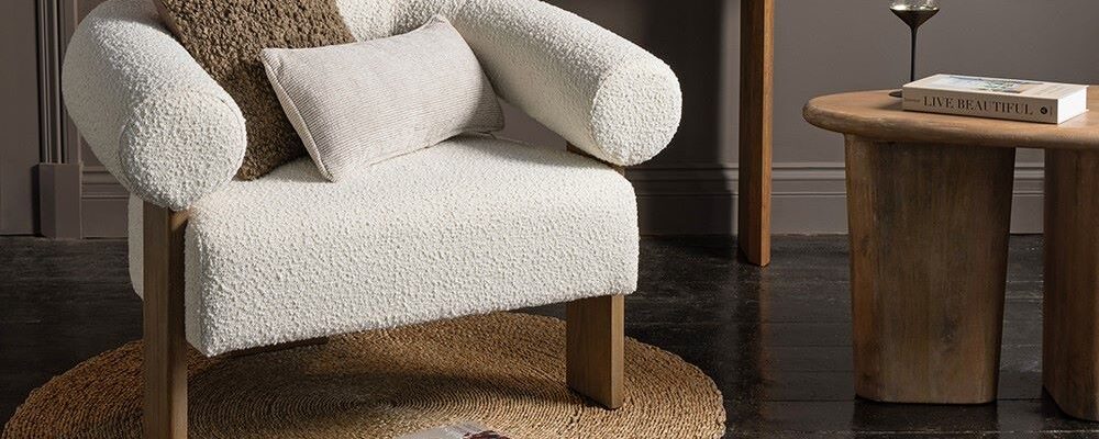 A cosy and stylish living space with a prominent white armchair taking centre stage. The chair has a unique curved design with rolled armrests and is upholstered in a soft, textured fabric. It sits on a round woven rug and is complemented by a wooden side table, a console table, and a collection of decorative items, including pillows, a glass, and books.