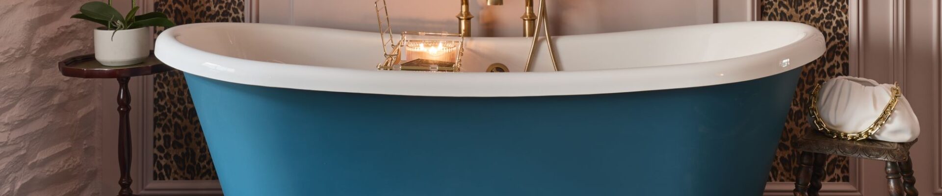 A freestanding bath with a roll top design. It has a white interior and a blue exterior. The bath is sitting on a carpeted floor in front of a wall with leopard print wallpaper. There is a gold faucet on the side of the bath.