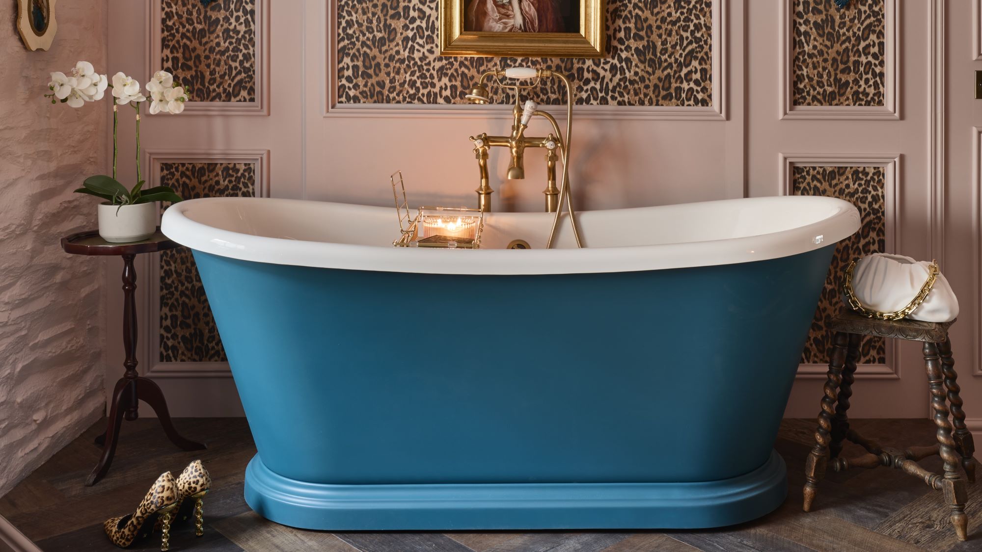 A freestanding bath with a roll top design. It has a white interior and a blue exterior. The bath is sitting on a carpeted floor in front of a wall with leopard print wallpaper. There is a gold faucet on the side of the bath.