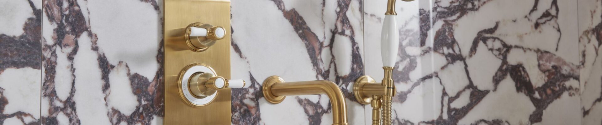 The image shows a luxurious bathroom with a Keswick Dual Function Concealed Shower System. The shower system has a brushed brass finish and features a thermostatic mixer valve with two ceramic grip levers for controlling temperature and flow. The shower head is a large, round overhead shower, and there is also a handheld shower with a flexible hose. The bathroom is also decorated with beautiful marble tiles and a freestanding bathtub.