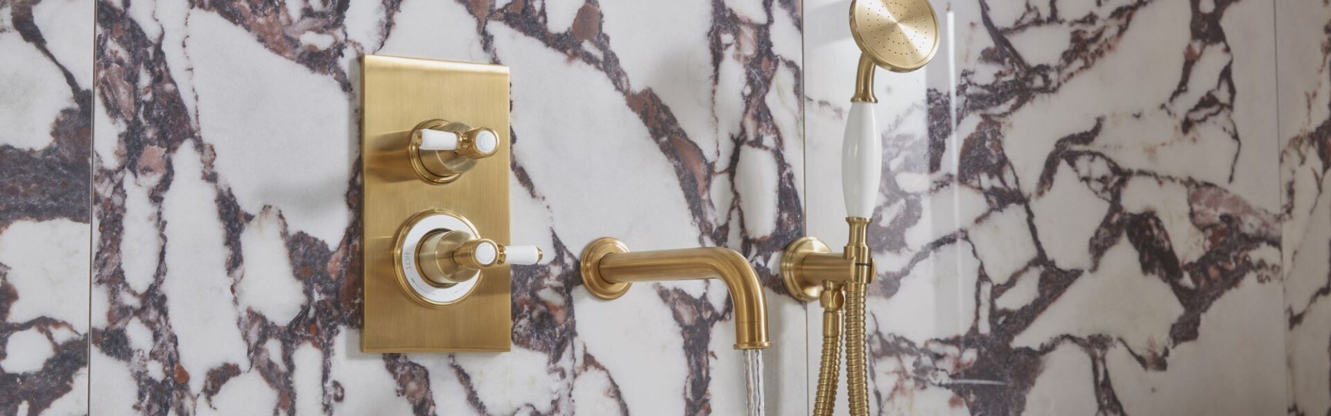 The image shows a luxurious bathroom with a Keswick Dual Function Concealed Shower System. The shower system has a brushed brass finish and features a thermostatic mixer valve with two ceramic grip levers for controlling temperature and flow. The shower head is a large, round overhead shower, and there is also a handheld shower with a flexible hose. The bathroom is also decorated with beautiful marble tiles and a freestanding bathtub.