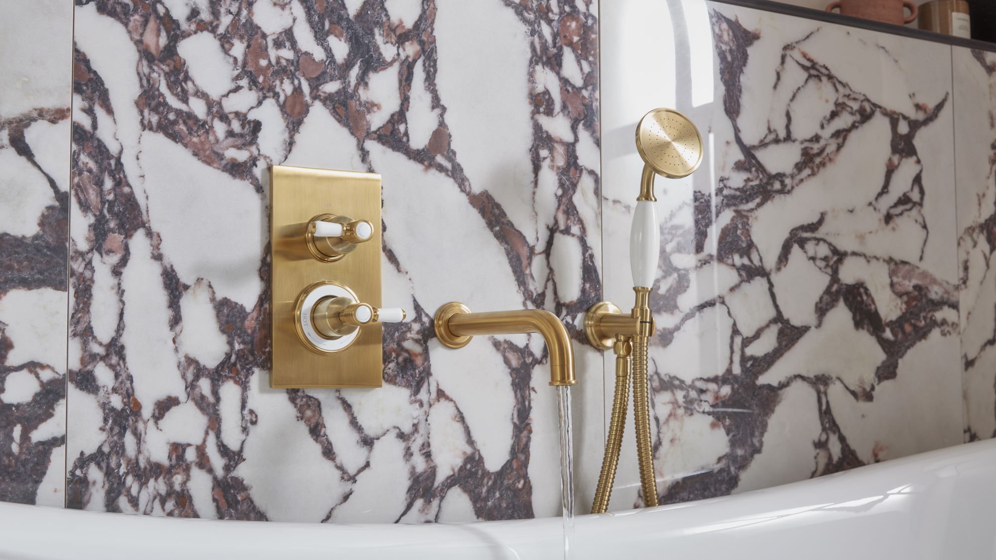 The image shows a luxurious bathroom with a Keswick Dual Function Concealed Shower System. The shower system has a brushed brass finish and features a thermostatic mixer valve with two ceramic grip levers for controlling temperature and flow. The shower head is a large, round overhead shower, and there is also a handheld shower with a flexible hose. The bathroom is also decorated with beautiful marble tiles and a freestanding bathtub.