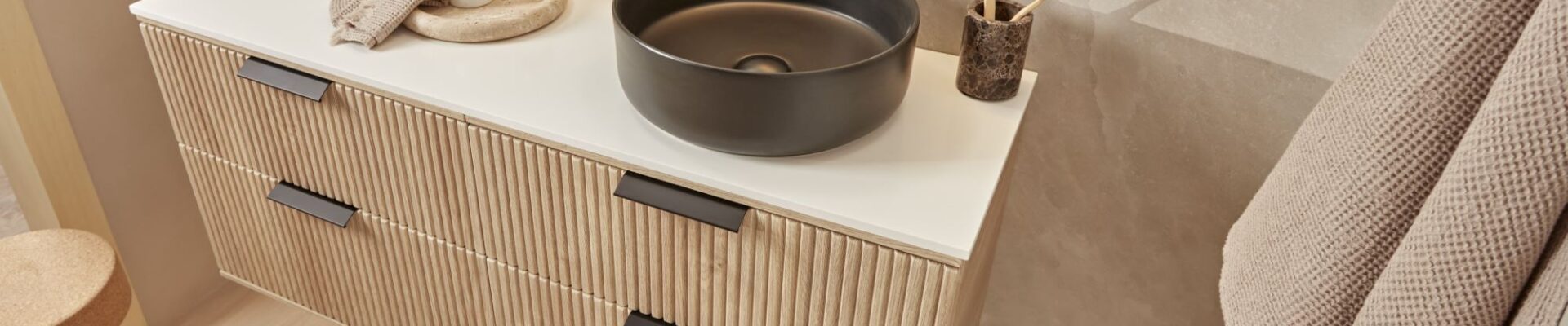 A modern bathroom vanity with a floating wooden cabinet and a black ceramic countertop. A round black sink sits on the countertop, with a black tap and soap dispenser next to it. A fluffy white towel hangs on the wall behind the vanity.
