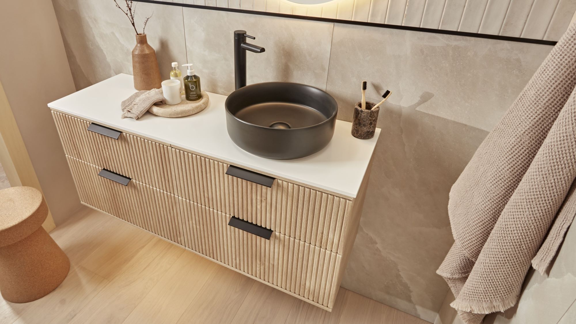 A modern bathroom vanity with a floating wooden cabinet and a black ceramic countertop. A round black sink sits on the countertop, with a black tap and soap dispenser next to it. A fluffy white towel hangs on the wall behind the vanity.