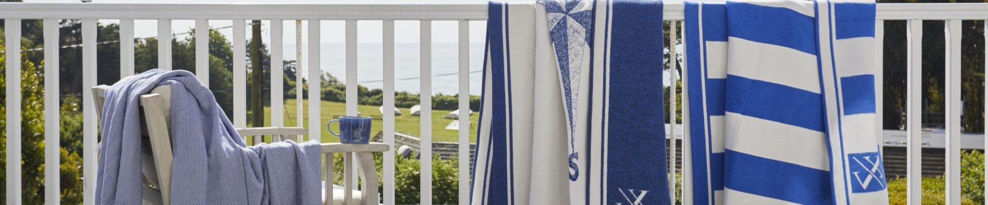 Blue and white striped blankets and throws draped over a white wooden balcony railing, with a blue blanket on a chair overlooking a coastal view.