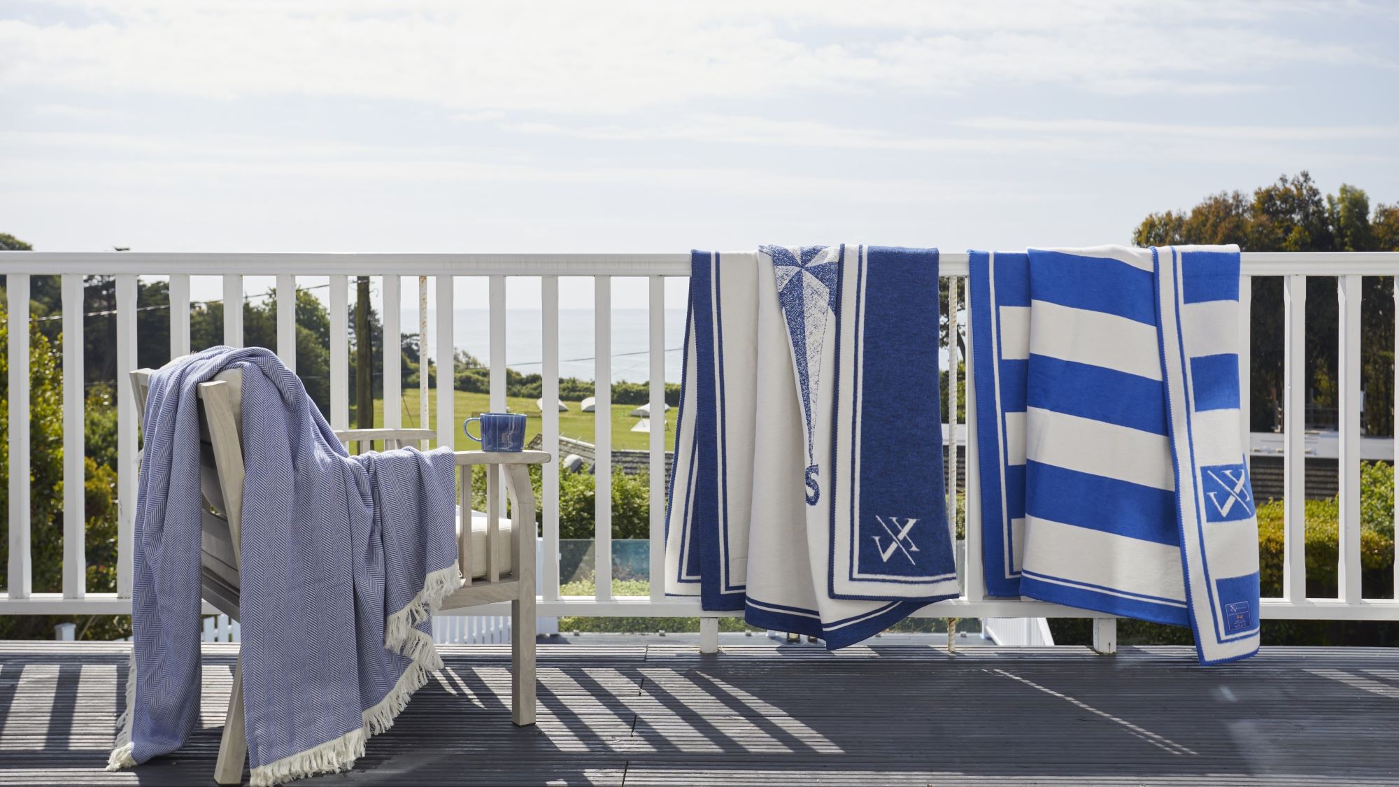 Blue and white striped blankets and throws draped over a white wooden balcony railing, with a blue blanket on a chair overlooking a coastal view.