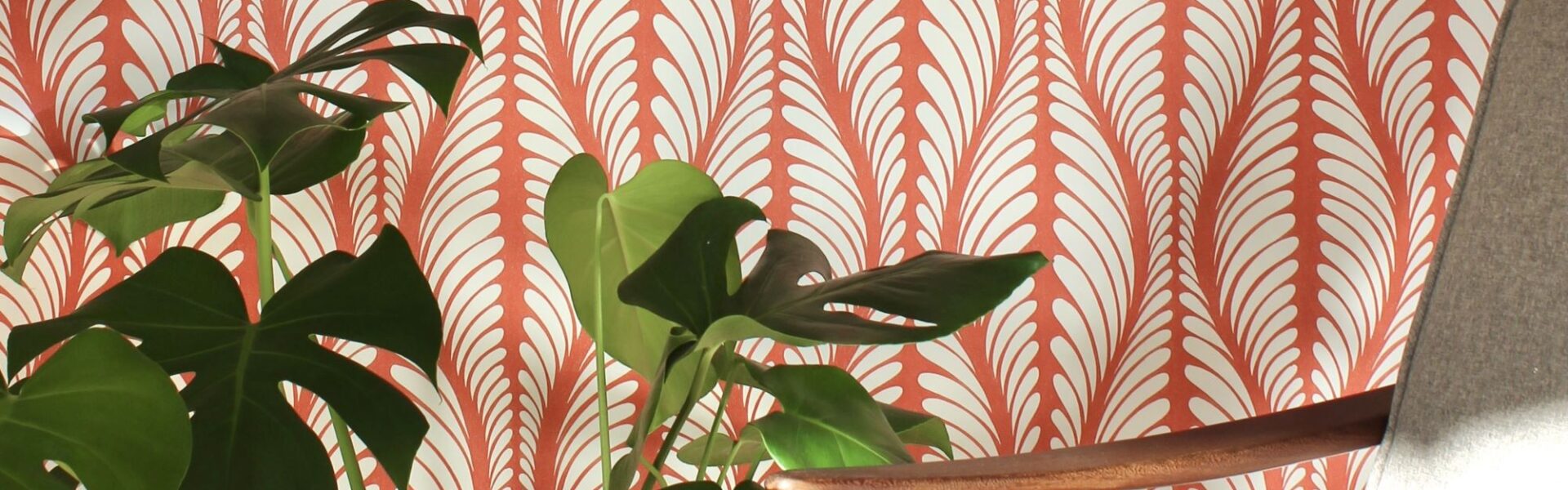 A cosy armchair sits in front of a wall covered in orange and white patterned wallpaper. A large houseplant with lush green leaves stands beside the chair.