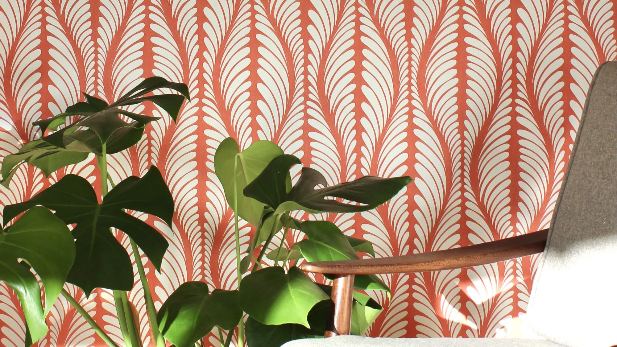 A cosy armchair sits in front of a wall covered in orange and white patterned wallpaper. A large houseplant with lush green leaves stands beside the chair.