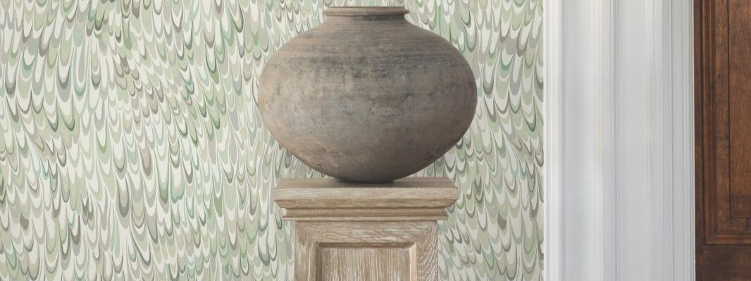 A large, earthy-toned clay pot sits on a wooden pedestal against a backdrop of a light green wallpaper with swirling, abstract feather-like patterns.
