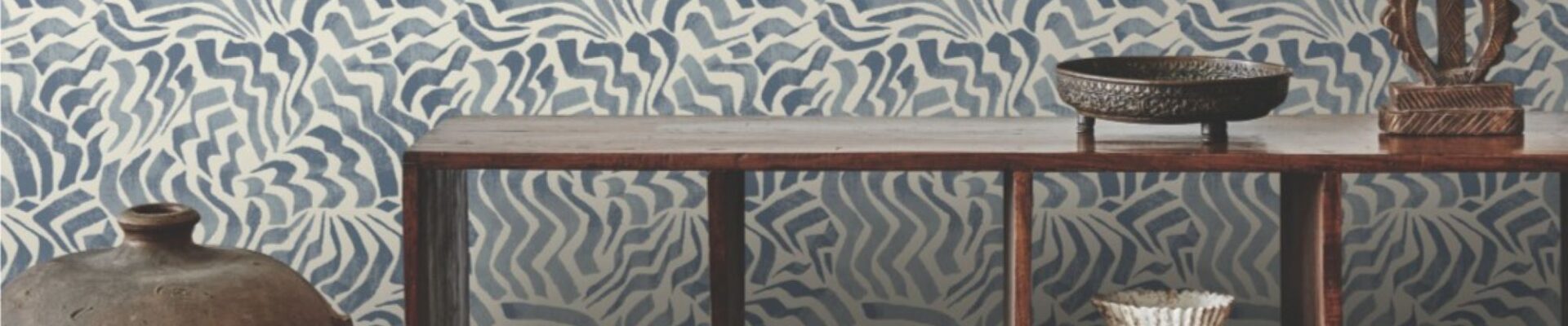 The image shows a room with a feature wall covered in a blue and white patterned wallpaper. The pattern is abstract and wavy, resembling stylised flowers or clouds. The wall is complemented by a wooden console table holding a collection of books, a vase, and a decorative bowl. A large ceramic jar stands to the left of the table, adding to the room's eclectic and cosy atmosphere.