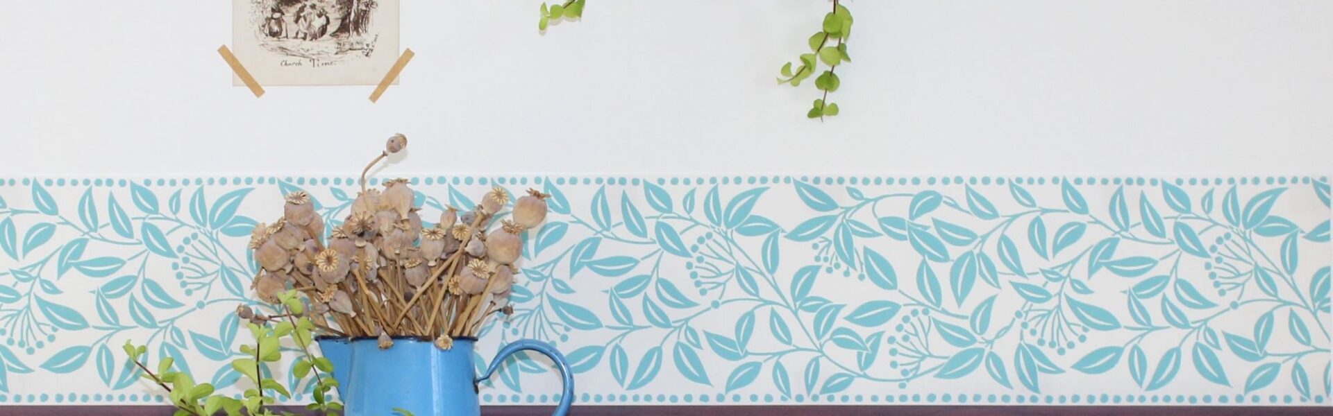 A room with a white wall and a wooden panelling at the bottom. There is a blue and white patterned border between the two. A picture frame with a black and white print hangs above the border. A vase with dried flowers sits on a table next to a small potted plant. A trailing plant hangs from a pot above the vase.