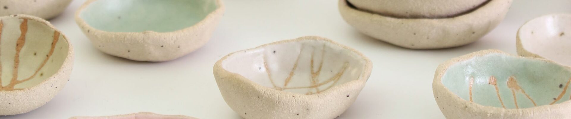 A collection of small, handmade ceramic bowls. They are arranged in groups on a white surface. The bowls vary in size and shape, and are decorated with different colours and patterns. Some bowls are plain white, while others are glazed in pastel colours such as pink, blue, and green. Some bowls have simple designs, such as lines or dots, while others have more intricate patterns.