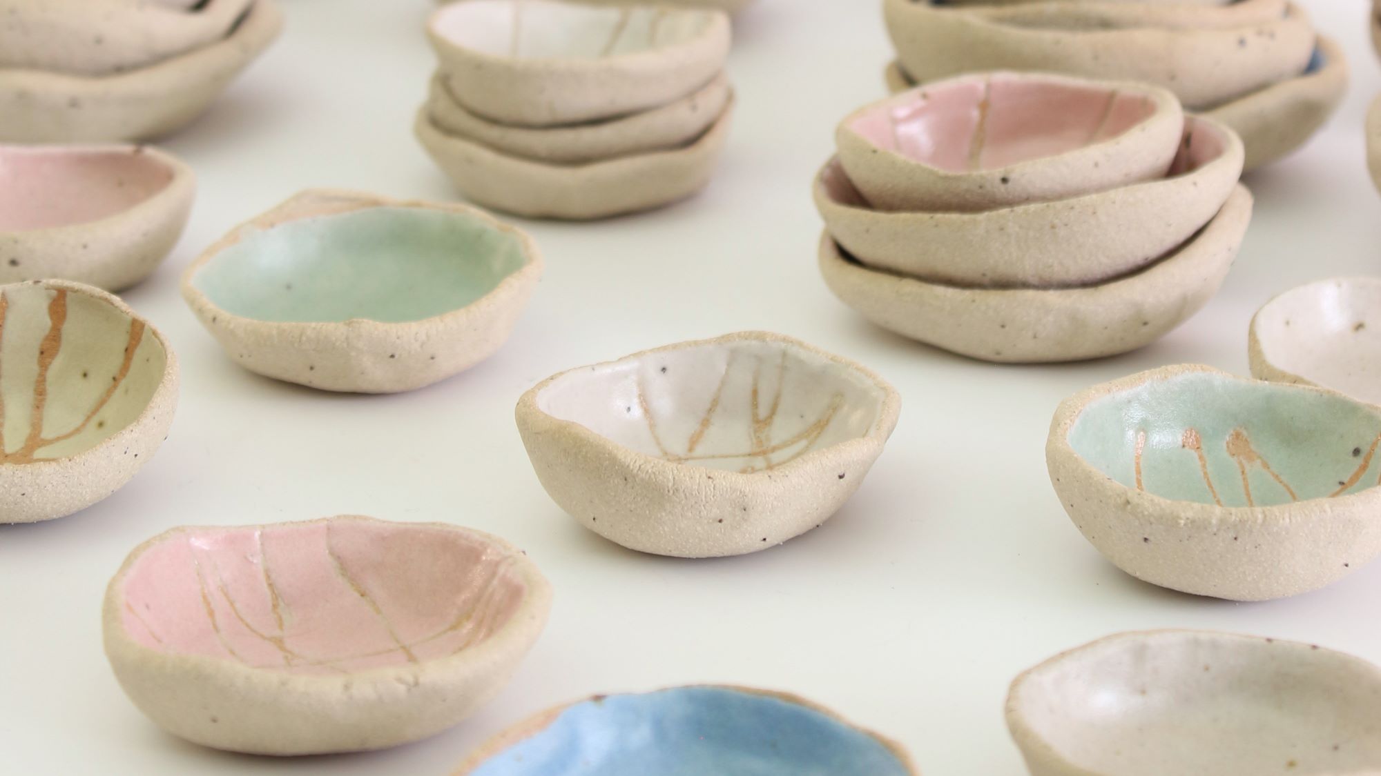 A collection of small, handmade ceramic bowls. They are arranged in groups on a white surface. The bowls vary in size and shape, and are decorated with different colours and patterns. Some bowls are plain white, while others are glazed in pastel colours such as pink, blue, and green. Some bowls have simple designs, such as lines or dots, while others have more intricate patterns.