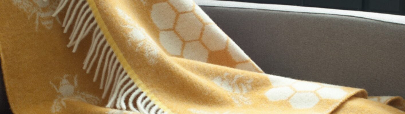The throw features a distinctive pattern of bees and honeycombs, woven into a soft, mustard yellow wool. The fringes add a touch of texture and style. It seems to be a cozy and visually appealing piece that would complement a variety of home decor styles.