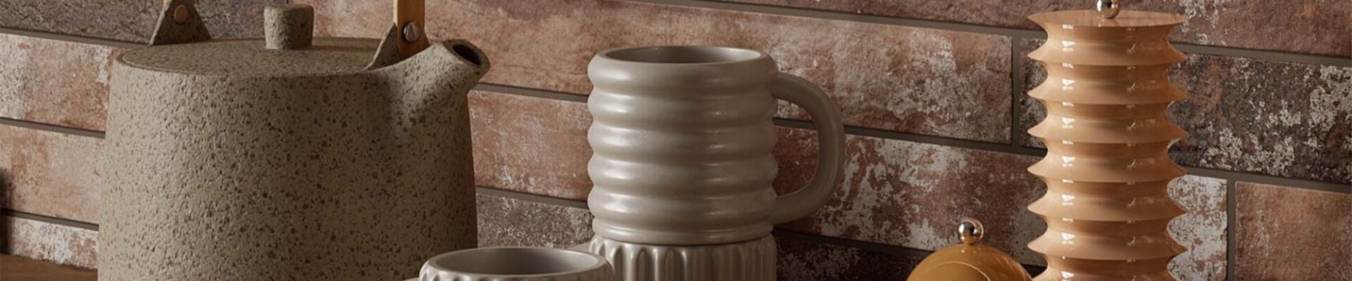 A kitchen countertop with a rustic brick wall backdrop. A wooden tray holds a teapot, a stack of mugs, and a set of salt and pepper shakers. The overall aesthetic is warm and inviting, with a focus on natural materials and earthy tones.
