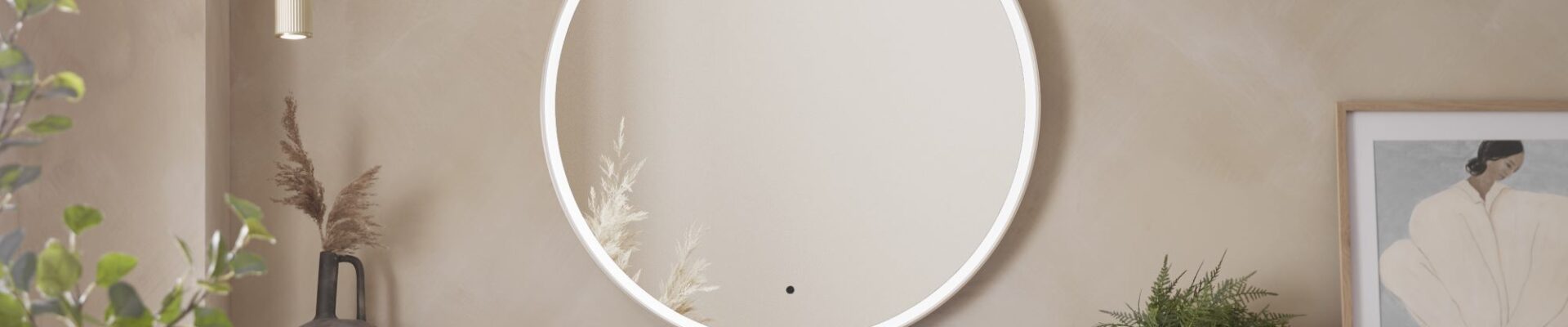 A round, illuminated mirror with a brass frame. It is hanging on a wall in a modern bathroom setting. There are also two gold pendant lights, a vase with dried flowers, a framed picture, and a potted plant.